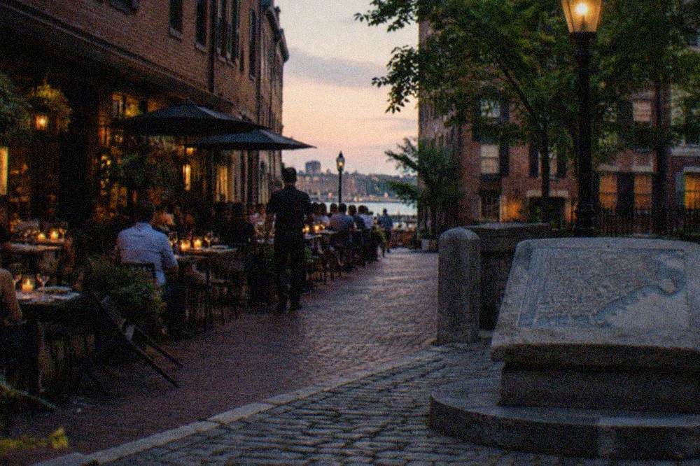 Crowded sidewalk tables outside Italian restaurants on a narrow North End street in the evening