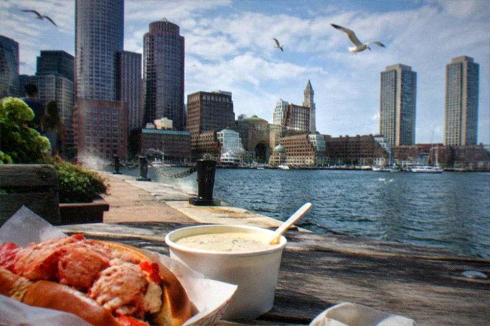 A split-top lobster roll in a paper basket beside a cup of chowder on a waterfront table