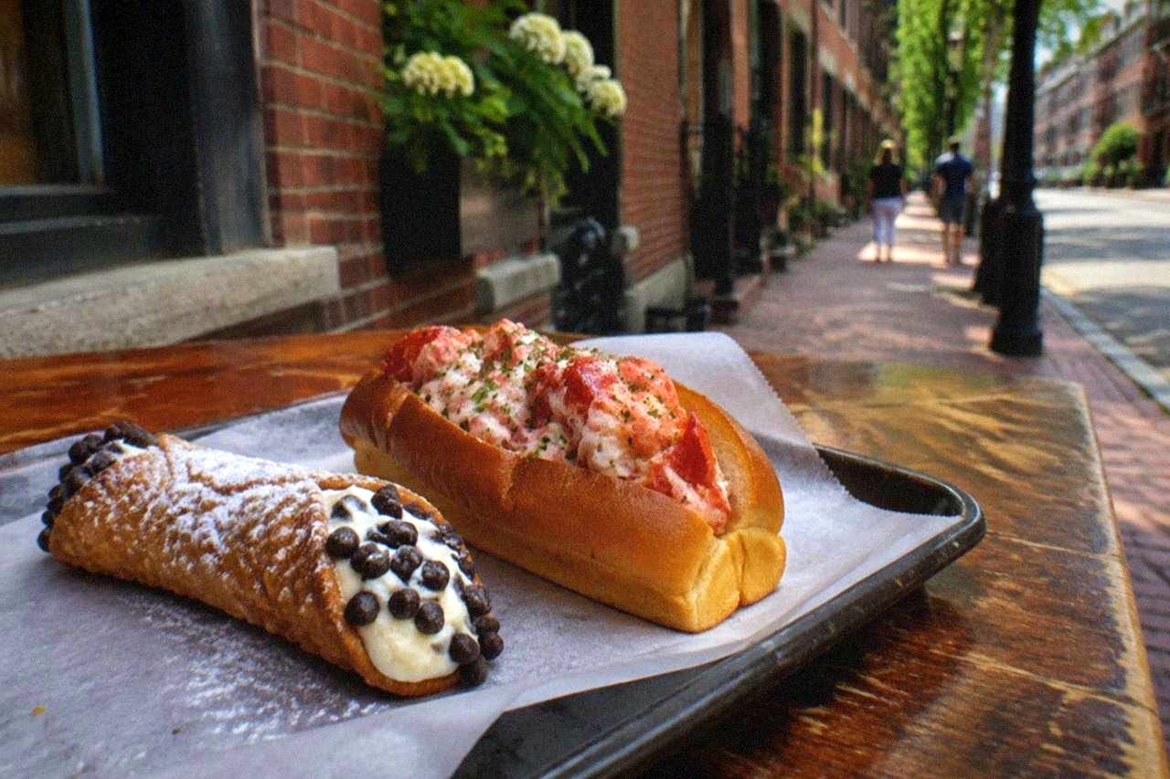 Close-up of a paper-lined tray holding a fresh cannoli and a hot lobster roll on a wooden counter