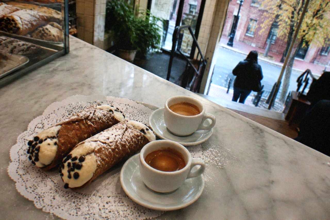 A pair of cannoli and two cups of espresso on a marble bakery counter in Bostons North End