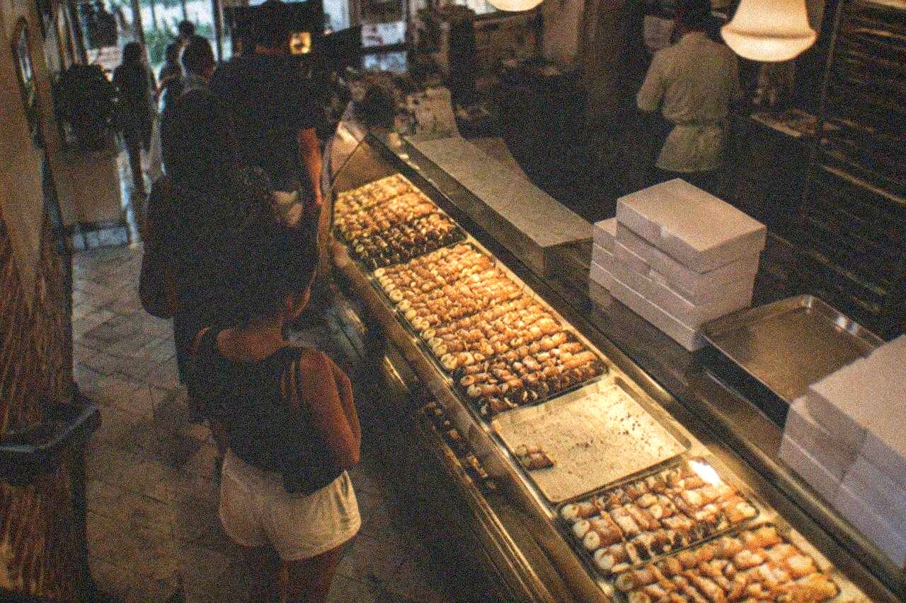 Bakery counter in a North End pastry shop with rows of cannoli under the lights