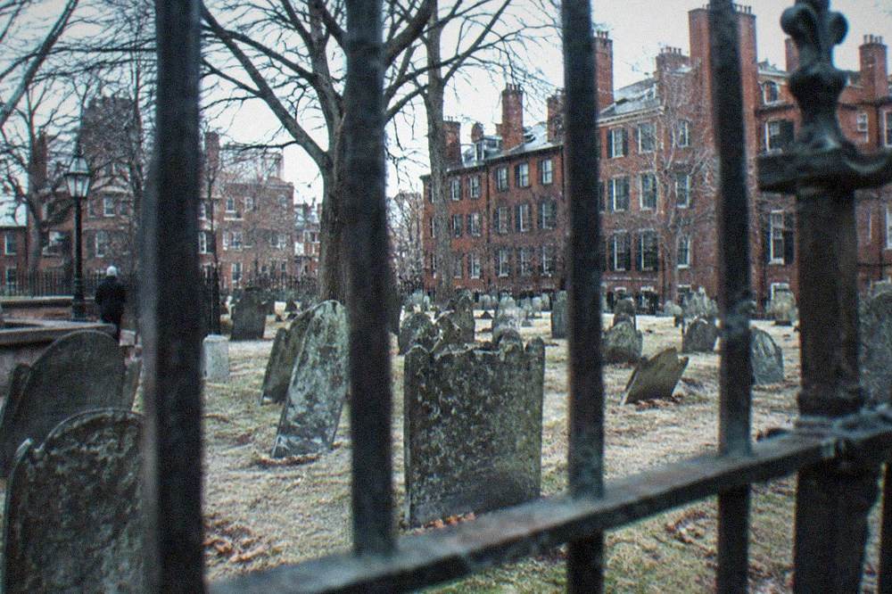 An old colonial era cemetery with weathered slate headstones and brick buildings visible beyond the fence