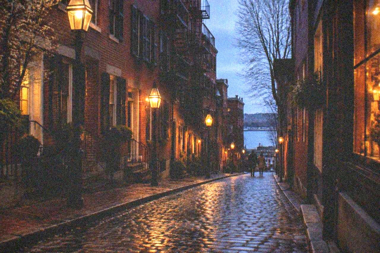 A narrow Boston street lined with old brick rowhouses and hanging shop signs in the evening