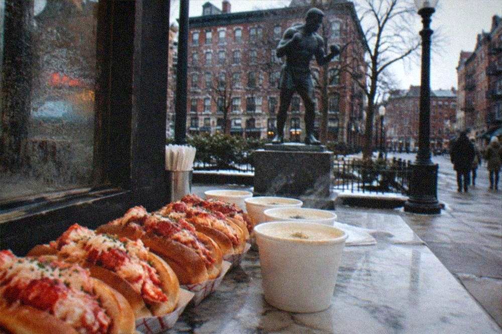 A restaurant chalkboard menu listing lobster roll prices beside a row of prepared rolls