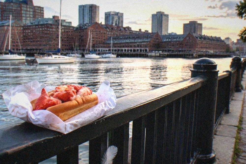 A harbor-side walking path with a lobster roll wrapped in paper held by a railing overlooking the water