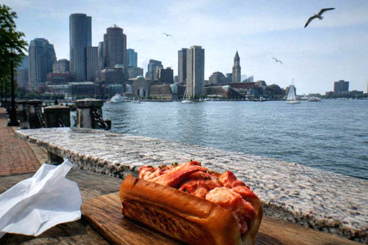 A toasted lobster roll with buttered meat served on a wooden board beside a harbor view