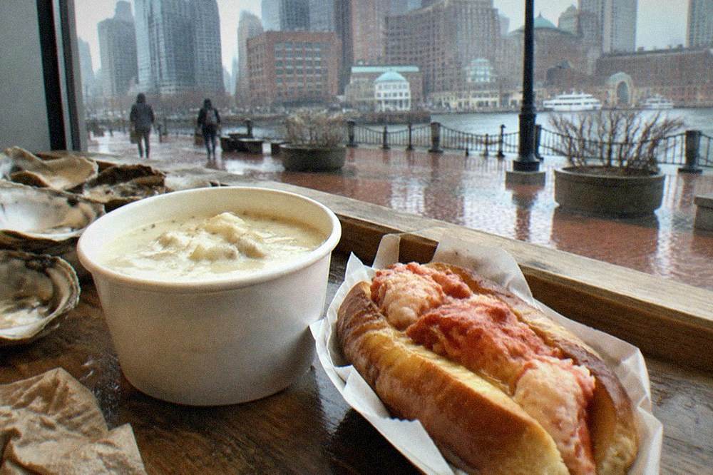 A cup of New England clam chowder beside a lobster roll on a wooden counter