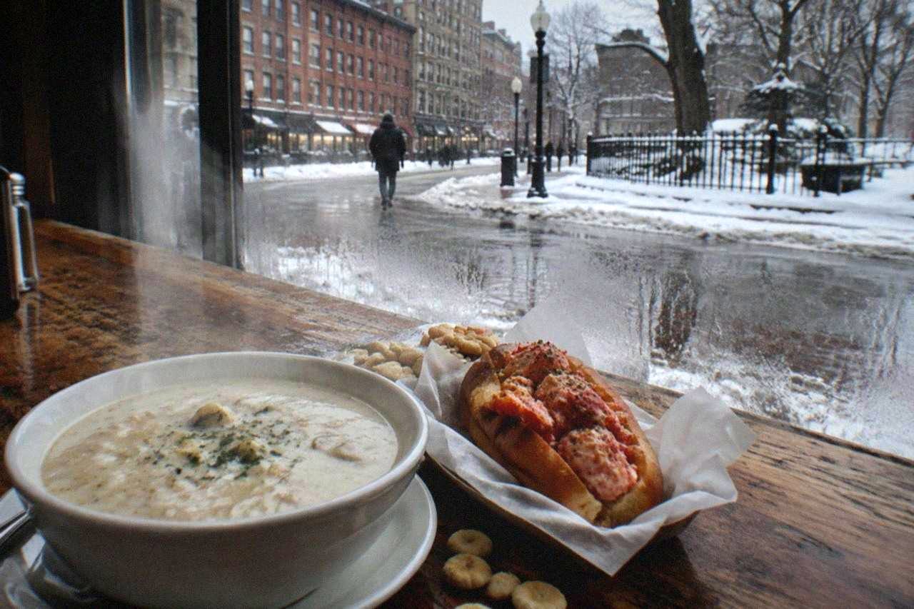 Bowl of creamy clam chowder beside a buttered lobster roll on a wooden restaurant table